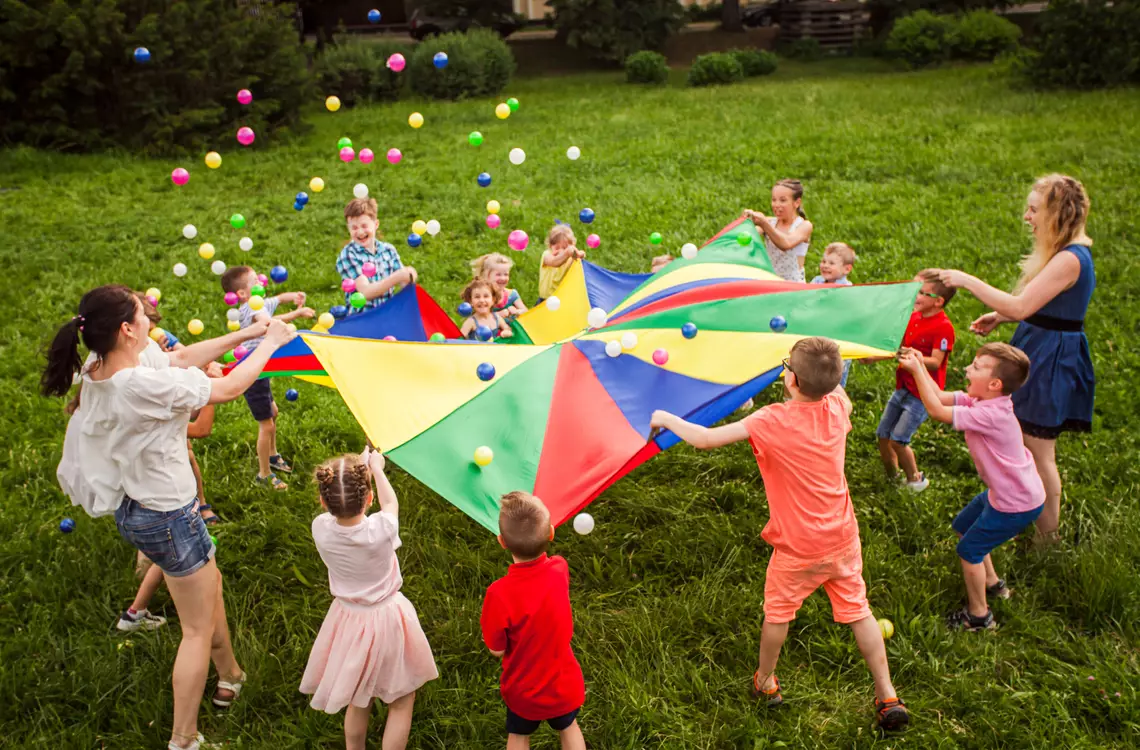 Des enfants de 6 à 10 ans et des adultes en train de jouer avec une toile de parachute en faisant sauter des petites balles dessus