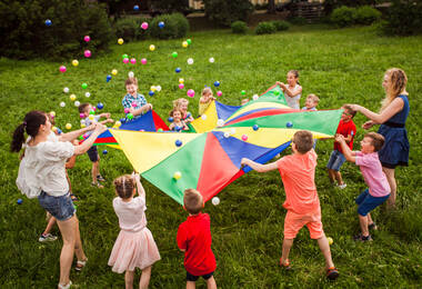 Des enfants de 6 à 10 ans et des adultes en train de jouer avec une toile de parachute en faisant sauter des petites balles dessus